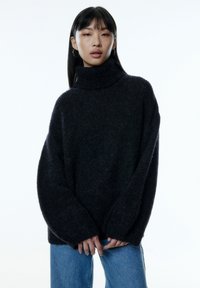 Pull oversized noir à col roulé en matière texturée, doté de manches longues et d'une coupe ample, associé à un jean bleu clair.
