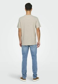 T-shirt beige chiaro a maniche corte, colletto rotondo, in cotone, abbinato a jeans azzurri e scarpe slip-on marroni, visto da dietro.