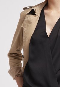 Beige trench coat dengan lengan digulung, menampilkan kerah lapel dan detail kancing, dilapisi di atas atasan hitam dengan leher v. Tekstur kain yang halus.