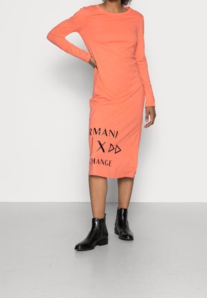 Femme portant une robe midi corail à manches longues avec un texte noir près de l'ourlet et des bottines noires, se tenant avec une main sur la hanche.