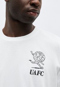 Κοντινό πλάνο ενός ατόμου που φορά λευκό πουκάμισο με μαύρο σχέδιο γραμμής ενός παγκόσμιου χάρτη με πόδια που κλωτσούν μια ποδοσφαιρική μπάλα και το κείμενο "UAFC".