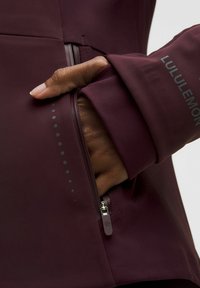 Burgundy idrottsjacka med dragkedjeficka, slät textur och diskret prickdetaljering längs ärmen. Logotyp synlig.