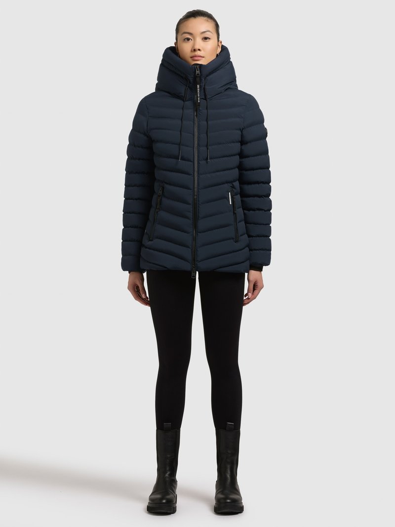 Tumesinine puffer-jope kõrge krae ja reguleeritava kapuutsiga, millel on horisontaalne tikand ja tõmblukuga taskud, sobitatud mustade leggingside ja saapadega.