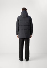 Black padded jacket with a hood, featuring horizontal quilted sections and a longer cut, paired with dark trousers and shoes.