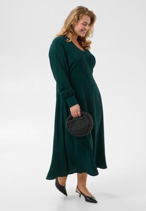 Femme en longue robe vert foncé tenant un sac à main tressé noir, portant des talons pointus noirs, souriante et regardant vers le bas.