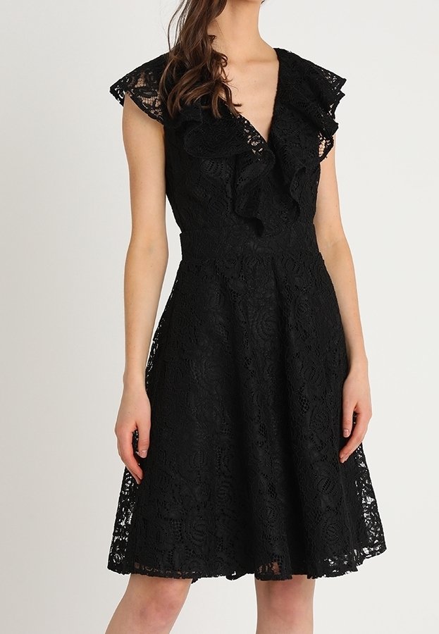 Robe en dentelle noire longueur genoux avec encolure en V volantée et manches gigot portée par une femme sur un fond clair uni.