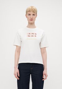 Hvit bomull T-skjorte med fet tekst "CANNES FRANKRIKE" i rødt, oransje og gult. Korte ermer og rund hals, brukt med mørke jeans.