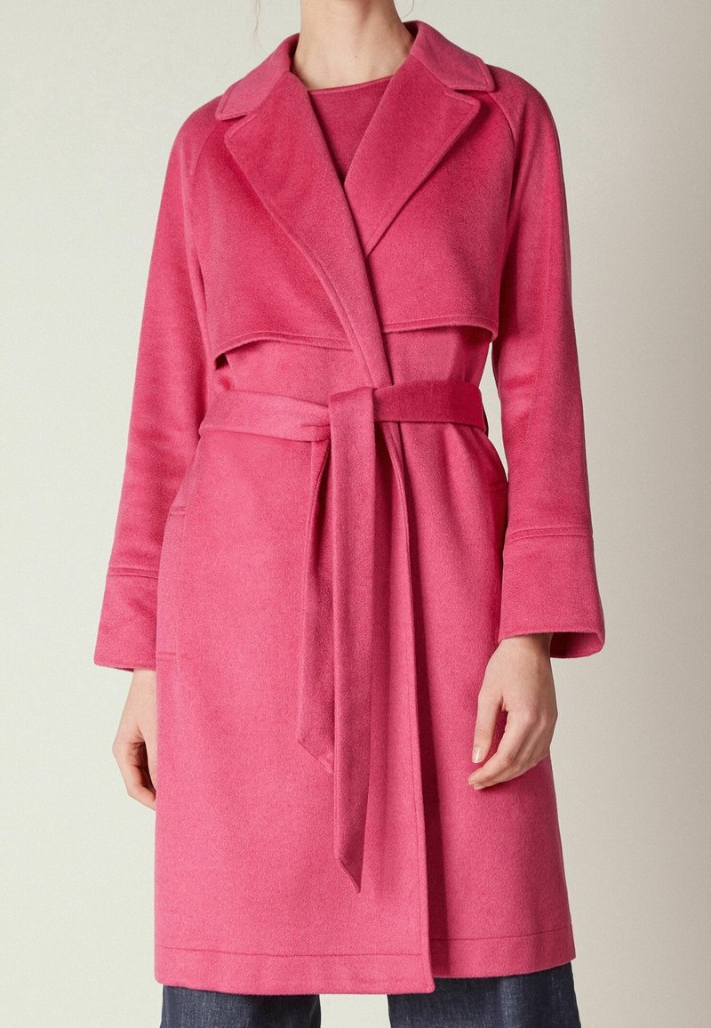 Manteau en laine rose avec col châle, taille ceinturée et poches latérales. Manches longues avec larges poignets, tissu texturé et longueur jusqu'aux genoux.