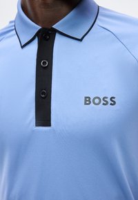 Κοντινό πλάνο ενός ατόμου που φορά ένα ανοιχτό μπλε πόλο με μαύρη λωρίδα με κουμπιά και λογότυπο "BOSS" στο στήθος.