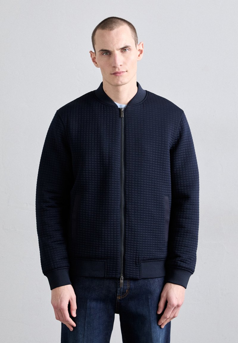 Bomber en tissu texturé bleu marine avec fermeture éclair, poignets et taille côtelés. Présente un motif à carreaux et des poches latérales, associé à un jean sombre.