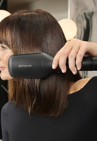 Czarna szczotka do prostowania włosów o eleganckim, prostokątnym kształcie. Posiada teksturowany uchwyt oraz widoczne logo na rączce. Włosy wydają się gładkie.