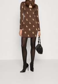 Robe à motifs marron à manches longues, avec un décolleté en forme de goutte. Portée avec des collants noirs et des escarpins noirs brillants, accompagnée d'un sac à main noir.