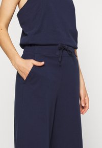 Vrouw draagt een marineblauwe mouwloze top en bijpassende wijde broek met trekkoord, hand in de zak, op witte achtergrond.