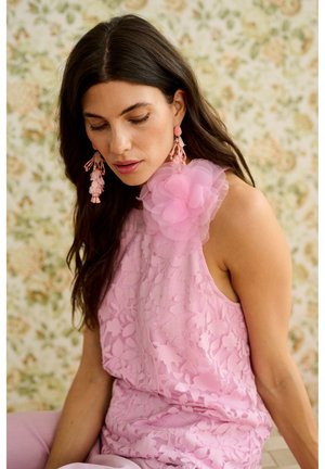 Kvinde med langt mørkt hår iført en ærmeløs pink blomsterkjole med en stor stofblomst på skulderen og matchende pink øreringe.