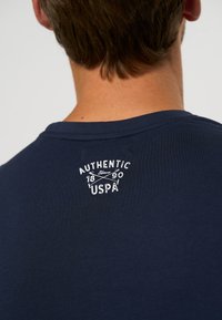 Marineblå bomull T-skjorte med rund hals og en hvit trykket logo på baksiden som sier "AUTHENTIC Siden 1890 USPA."