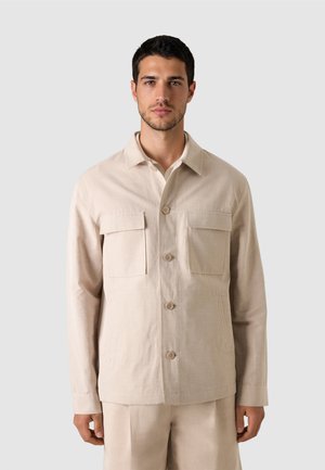 Ein Mann steht mit Blick nach vorne und trägt eine beige geknöpfte Hemdjacke mit zwei Brusttaschen sowie passende Hosen vor einem schlichten Hintergrund.