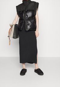 Osoba w czarnej kamizelce bez rękawów, czarnej spódnicy do kostek z bocznym rozcięciem, czarnych butach wsuwanych, niosąca torebkę w dwukolorze szaro-czarnym.