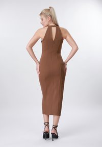 Γυναίκα με ξανθό κοτσίδι που φοράει καφέ εφαρμοστό μίντι φόρεμα χωρίς μανίκια και μαύρα σανδάλια με ψηλό τακούνι, στέκεται με τα χέρια στη μέση, με την πλάτη στραμμένη προς τα εμπρός.