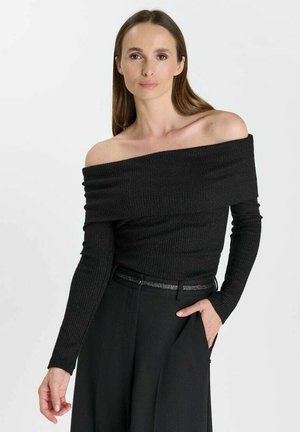Donna con lunghi capelli castani che indossa un top nero con spalle scoperte e maniche lunghe e pantaloni neri, con una mano in tasca, su sfondo bianco.
