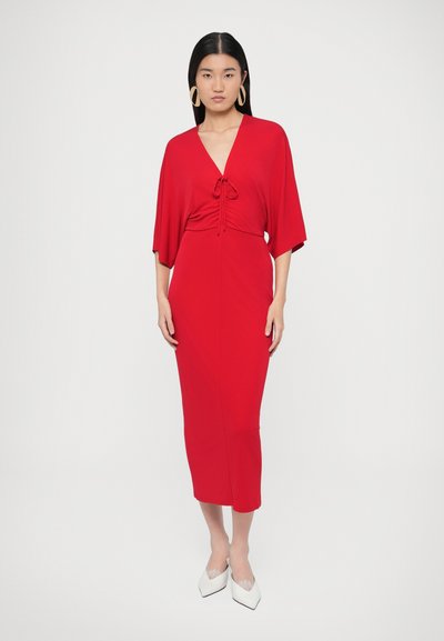 Sarkana ceļa garuma kleita no mīksta auduma ar V veida izgriezumu un jostas detaļu viduklī. Kleitai ir platas piedurknes un pieguloša siluets; valkāta ar baltām augstpapēžu kurpēm.