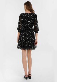 Robe noire transparente à pois blancs, manches courtes bouffantes, taille élastique et ourlet à volants. Dotée d'un col rond et d'une fermeture à boutons au dos.