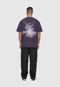 Lilla bomulds t-shirt med et stort grafisk motiv af en vinget figur og teksten "Celestial Society" på ryggen. Løs pasform, korte ærmer.
