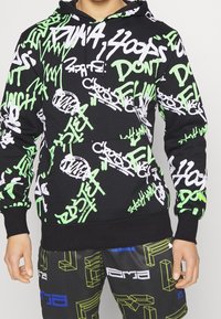 Felpa nera con motivi di testo in stile graffiti verde e bianco; presenta un cappuccio con cordini e polsini a coste; abbinata a pantaloni corti con grafica.