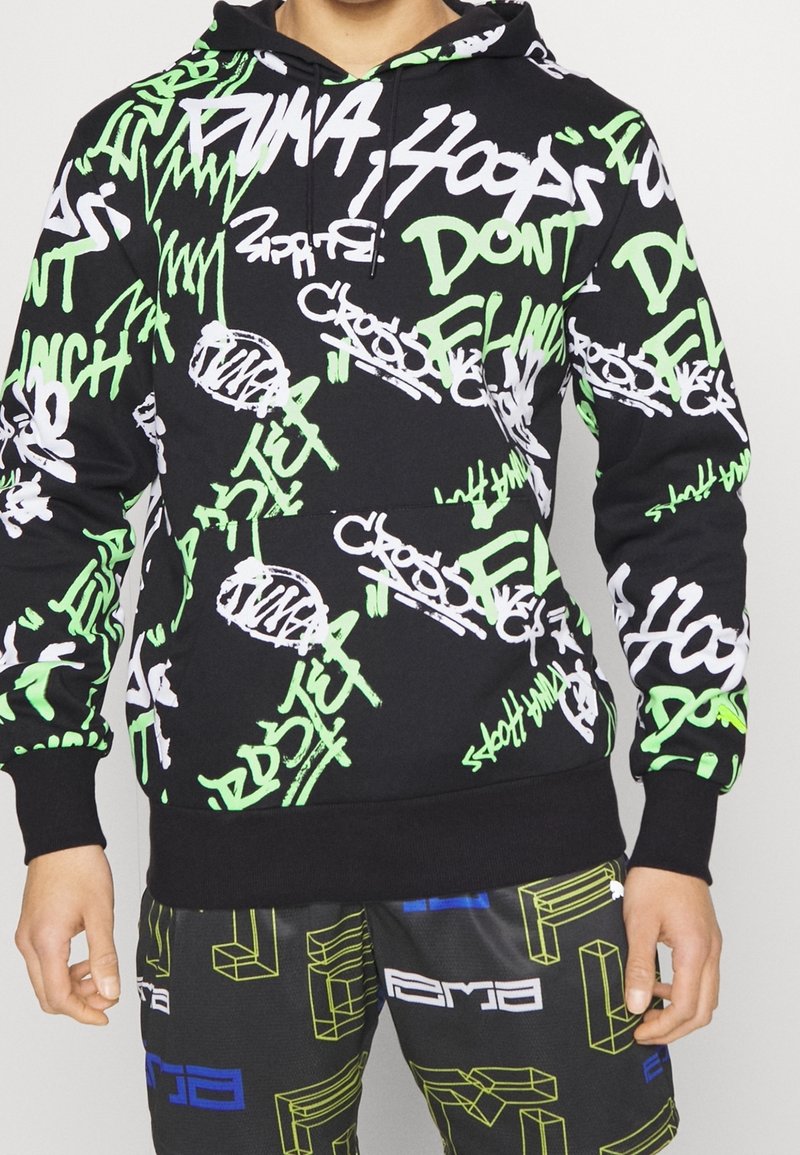 Felpa nera con motivi di testo in stile graffiti verde e bianco; presenta un cappuccio con cordini e polsini a coste; abbinata a pantaloni corti con grafica.