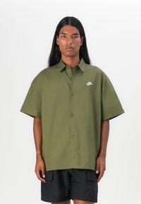 Olive zaļš īsām piedurknēm krekls ar pogu aizdari un mazu baltu Nike logotipu kreisajā krūtī. Gluda tekstūra un brīvs piegriezums.