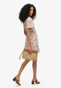 Clutch de tejido beige con detalles de cuero marrón y borlas, con forma rectangular. Modelo lleva un vestido de pata de gallo en tonos pastel y tacones.