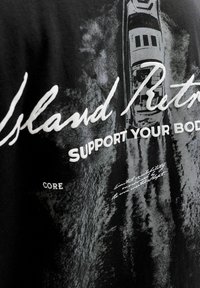 Tissu noir avec texte blanc "Island Retro" et "SUPPORT YOUR BODY" sur une vue aérienne en niveaux de gris d'un bateau sur l'eau.