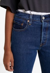 Pantalones vaqueros de mezclilla azul oscuro con una cintura de altura media, remaches de cobre y costuras contrastantes. Presentan un bolsillo frontal y cierre de botón.