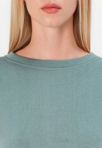 Cămașă cu guler rotund dintr-un material moale de culoare teal; prezintă o textură subtilă și un finisaj neted, cu un design simplu al gulerului și fără tipare sau accente vizibile.