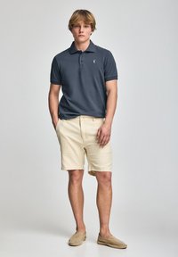 Camisa polo azul marino con tres botones y logo blanco, combinada con pantalones cortos de un beige claro y zapatos de lona beige. Fondo simple.