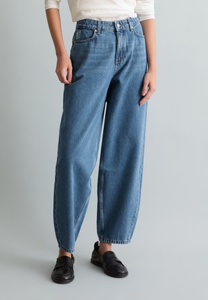 Person wearing high-waisted loose-fit blue jeans, white long-sleeve top, and black slip-on shoes, standing against plain light background.