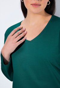 Tyrkysový dlouhý rukáv s výstřihem do V. Hladká tkanina, uvolněný střih a viditelné švy poblíž výstřihu. Model jemně hladí klíční kost.