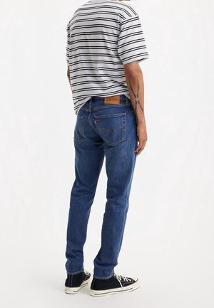 Niebieskie dżinsy o slim fit, w połączeniu z szaro-czarną koszulą w paski. Z tyłu znajduje się skórzana łatka. Widoczne czarne trampki.