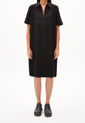 Schwarzes Kurzarmkleid mit Reißverschluss vorne und Kragen. Glatte Textur, knielange Silhouette. Kombiniert mit schwarzen klobigen Absatzclogs.