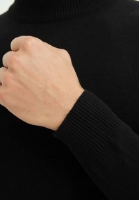 Pull marin noir, détail de poignet côtelé, texture douce, vue en gros plan d'un poing serré et d'un poignet, mettant en avant le matériau et le design.