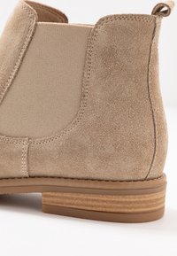 Stivaletto alla caviglia in camoscio beige con pannelli elastici laterali, una texture liscia e un tacco impilato marrone chiaro. Presenta cuciture visibili lungo le cuciture.
