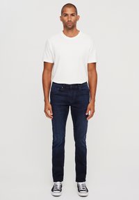 Muška odjeća koja uključuje bijelu majicu s okruglim izrezom i tamnoplave uske traperice, uparene s crno-bijelim tenisicama, stoji na neutralnoj pozadini.