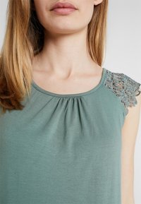 Grön ärmlös topp i mjukt tyg, med gathered-detaljer vid halsringningen och intrikata spetstillägg på axeln.