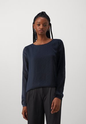 Blouse à manches longues bleu marine avec des pois texturés, col rond et poignets froncés. Associée à un pantalon tailleur gris foncé.