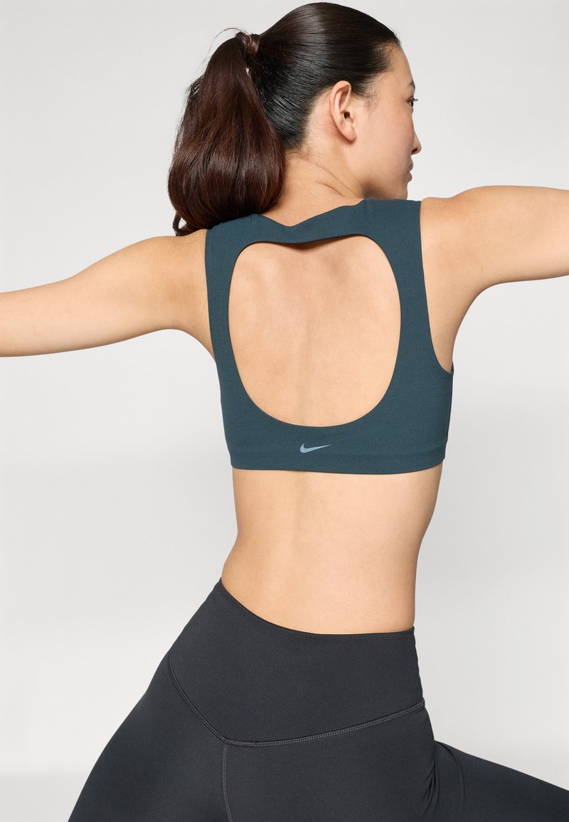Sportbeha in donker teal met een breed open rugontwerp. Heeft een subtiel logo in het midden en een gladde, rekbare stof.