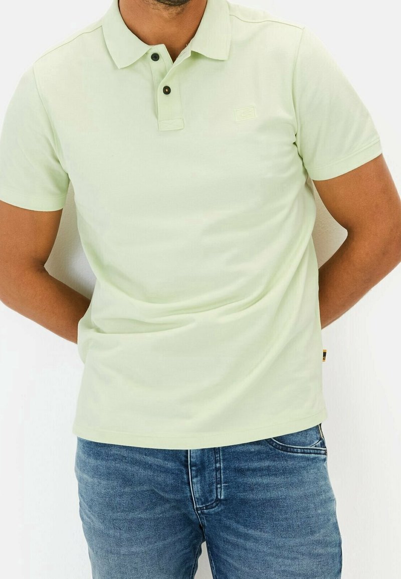 Homme portant un polo vert clair à manches courtes et un jean bleu avec les mains dans le dos sur un fond uni.