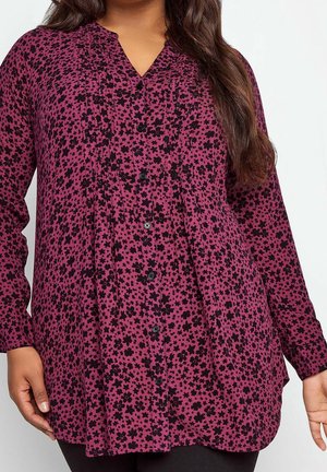 Woman wearing long-sleeve magenta tunic with black floral and star patterns, buttoned front, and loose fit, showing upper body and arms.