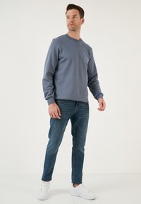 Felpa grigia in cotone a maniche lunghe con polsini a coste, abbinata a jeans in denim blu e sneaker bianche, su uno sfondo neutro.
