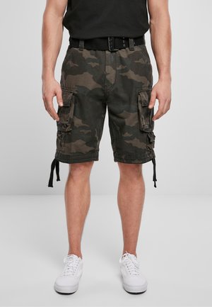 Homme portant un t-shirt noir, un short cargo camouflage avec poches latérales, une ceinture noire et des baskets blanches, debout devant un fond uni.