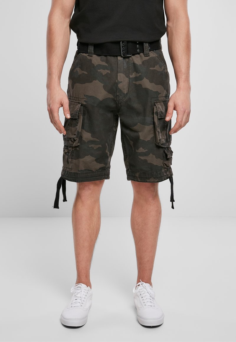 Homme portant un t-shirt noir, un short cargo camouflage avec poches latérales, une ceinture noire et des baskets blanches, debout devant un fond uni.