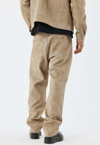 Pantalones marrones claros de tela texturizada; corte suelto con detalles de costura en el bolsillo trasero. Combinados con zapatos negros.